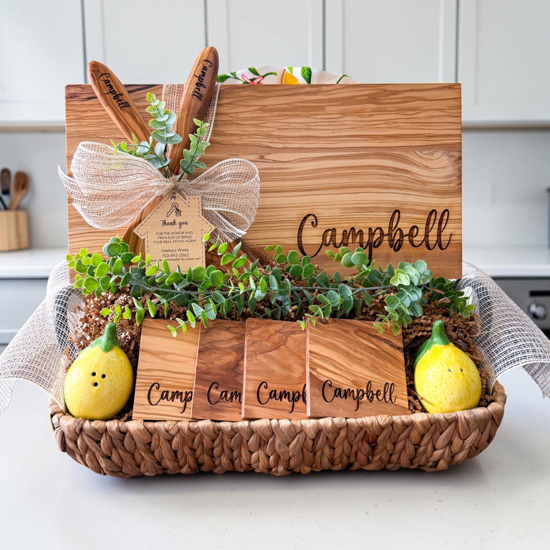 Luxury personalized olive wood realtor closing gift basket featuring engraved cutting board, custom coasters, olive wood serving spoons, greenery, and house-shaped thank you tag for home buyers, housewarming gifts, and real estate client appreciation gifts.
