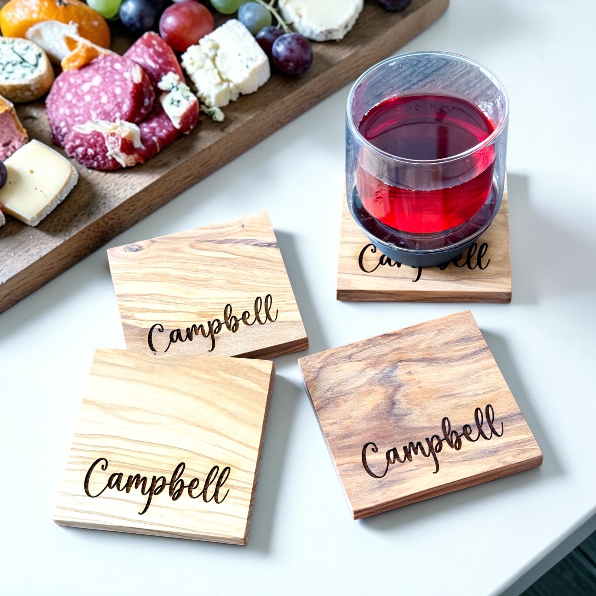 Personalized olive wood coasters engraved with family name beside charcuterie board and wine glass, luxury housewarming gift for new homeowners, realtor closing gift, custom kitchen decor, and thoughtful real estate client gift.