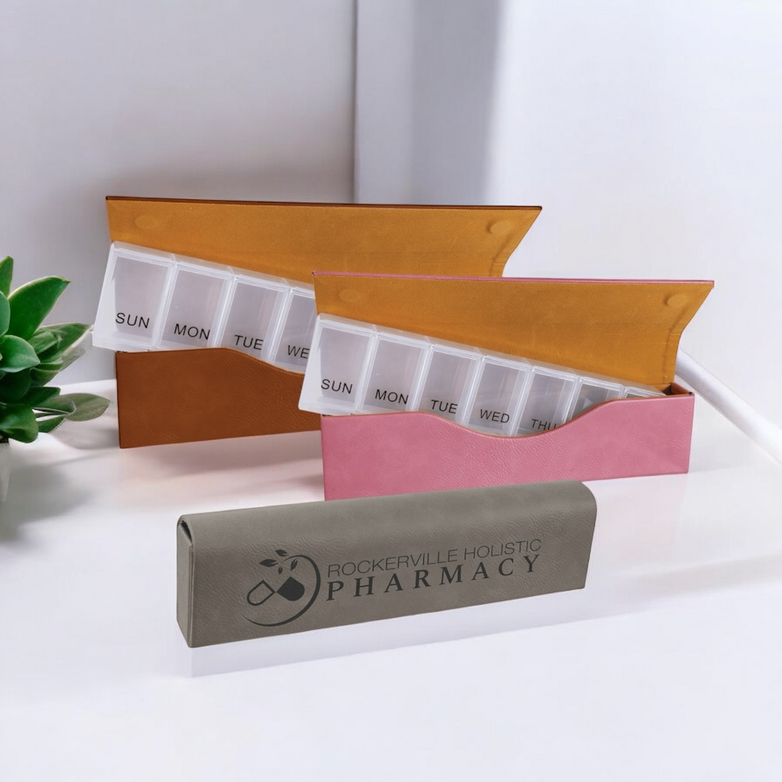 Custom Laserable Leatherette Pill Box with Weekly Organizer, featuring a sleek design with seven large compartments labeled for each day of the week, displayed in pink, rawhide, and gray leatherette cases with a magnetic closure. Ideal for corporate branding, travel, and personal use.