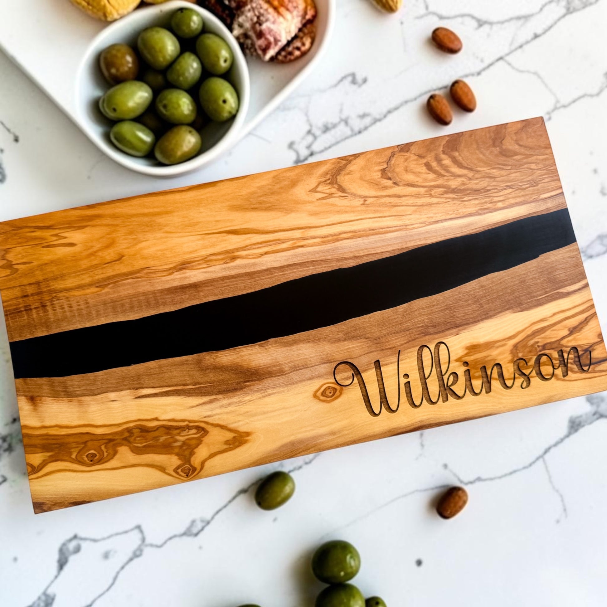 Luxury realtor closing gift featuring personalized olive wood and black resin charcuterie board engraved with Wilkinson family name styled with olives and appetizers for home buyers