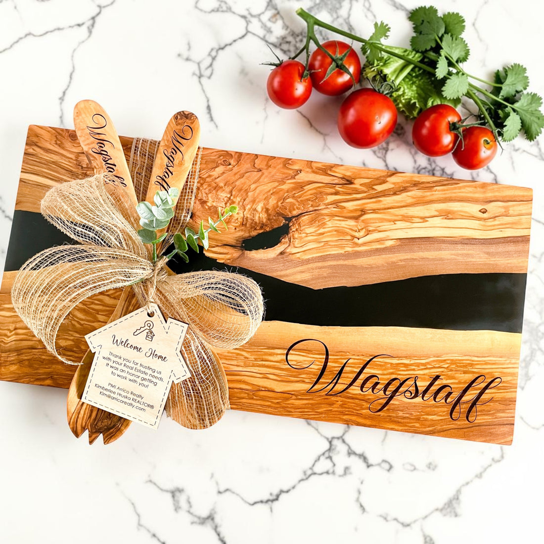 Personalized olive wood charcuterie board with black resin river detail, engraved “Cole & Kaylin” design, rustic burlap bow, and custom realtor thank-you tag styled with fresh tomatoes and herbs on a light neutral background. Luxury real estate closing gift and personalized kitchen gift for new homeowners.