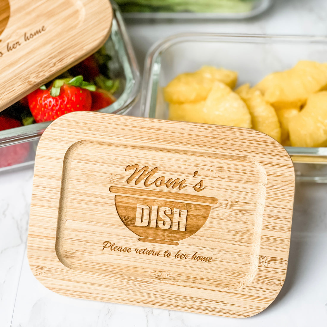 Personalized glass food storage container set with engraved bamboo lids featuring “Mom’s Dish – Please Return to Her Home” design. Elegant reusable kitchen storage containers styled with fresh fruit and vegetables, perfect for thoughtful realtor closing gifts, housewarming gifts, and custom kitchen décor.