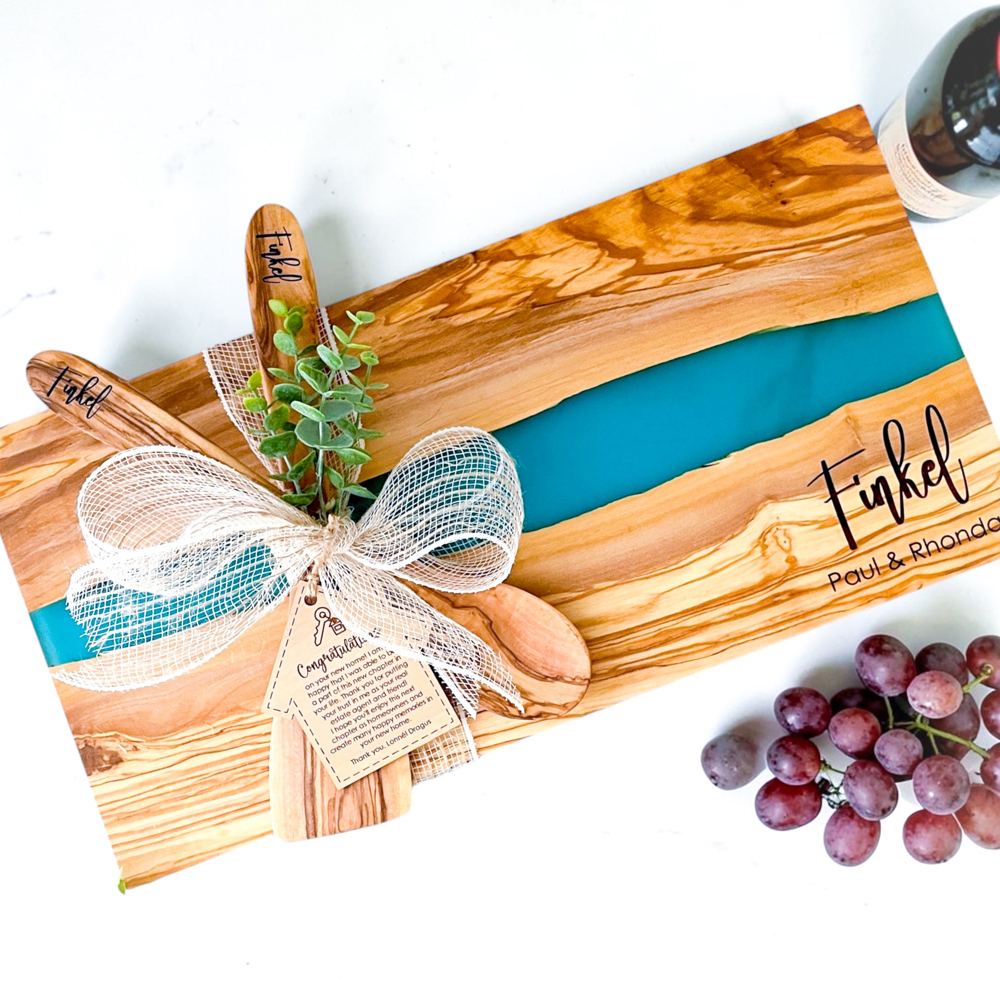 Luxury personalized blue river resin and olive wood charcuterie board with engraved names, wooden serving spoons, and custom realtor gift tag designed as an upscale closing gift for home buyers and housewarming celebrations