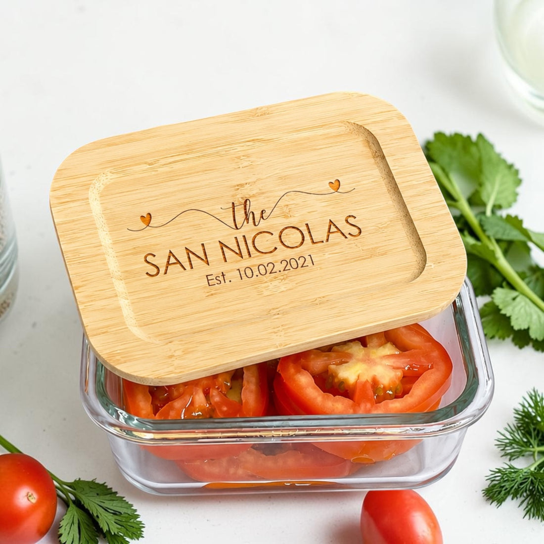 Personalized glass food storage container set with engraved bamboo lids featuring custom family name and established date. Elegant reusable kitchen storage containers styled with fresh vegetables and tomatoes for luxury realtor closing gifts, personalized housewarming gifts, and modern kitchen organization.