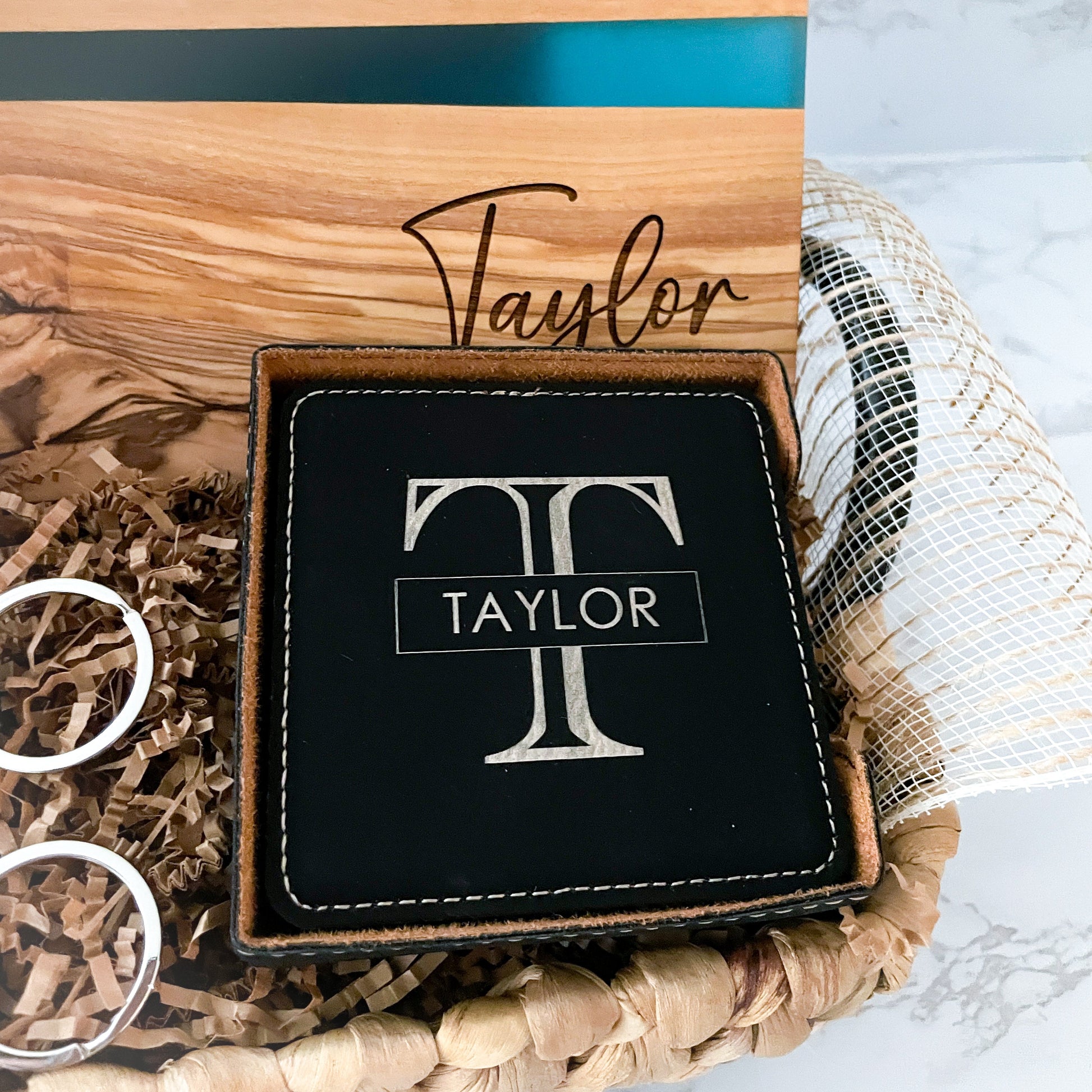 Personalized black leatherette coaster set with engraved monogram and family name displayed in a luxury realtor closing gift basket with olive wood serving board