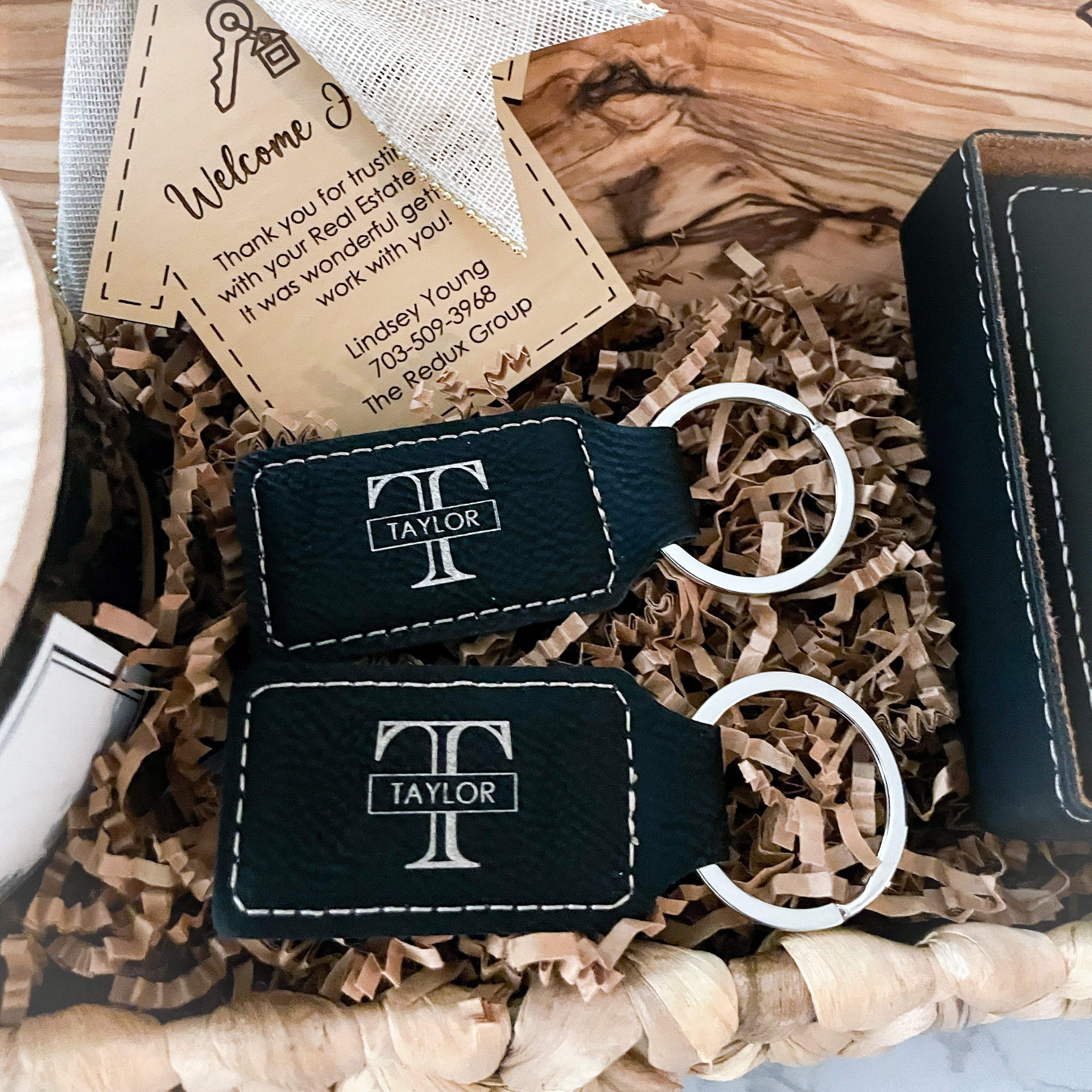 Personalized black leatherette keychains with engraved monogram logo displayed in a luxury realtor closing gift basket alongside olive wood serving board accessories