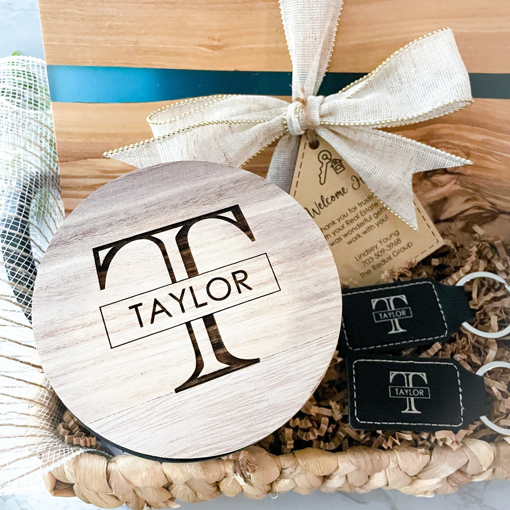 Personalized monogram candle displayed in a luxury realtor closing gift basket with engraved leatherette keychains and blue resin olive wood serving board accessories
