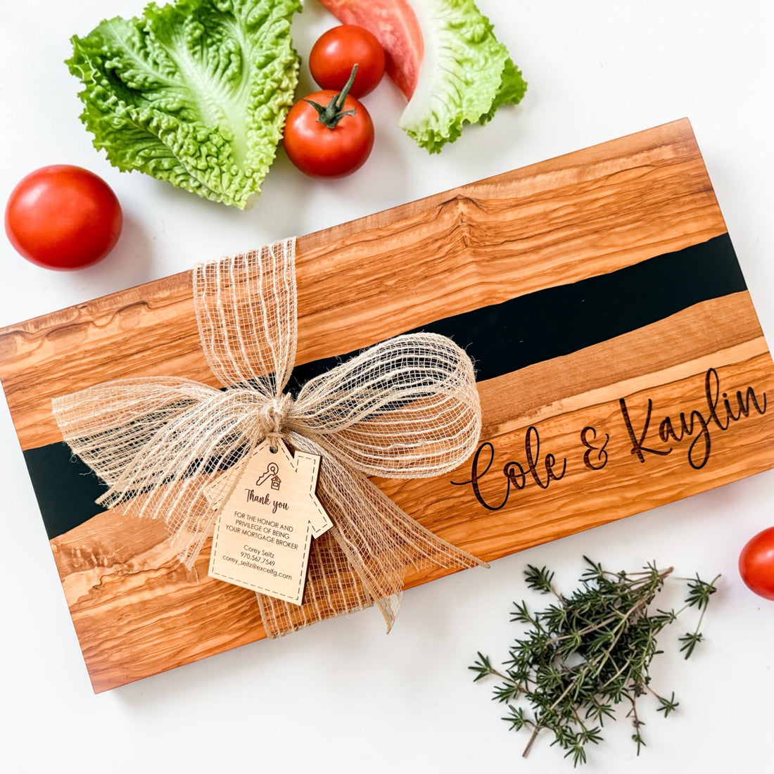 Personalized olive wood charcuterie board with black resin river detail, engraved “Cole & Kaylin” design, rustic burlap bow, and custom realtor thank-you tag styled with fresh tomatoes and herbs on a light neutral background. Luxury real estate closing gift and personalized kitchen gift for new homeowners.