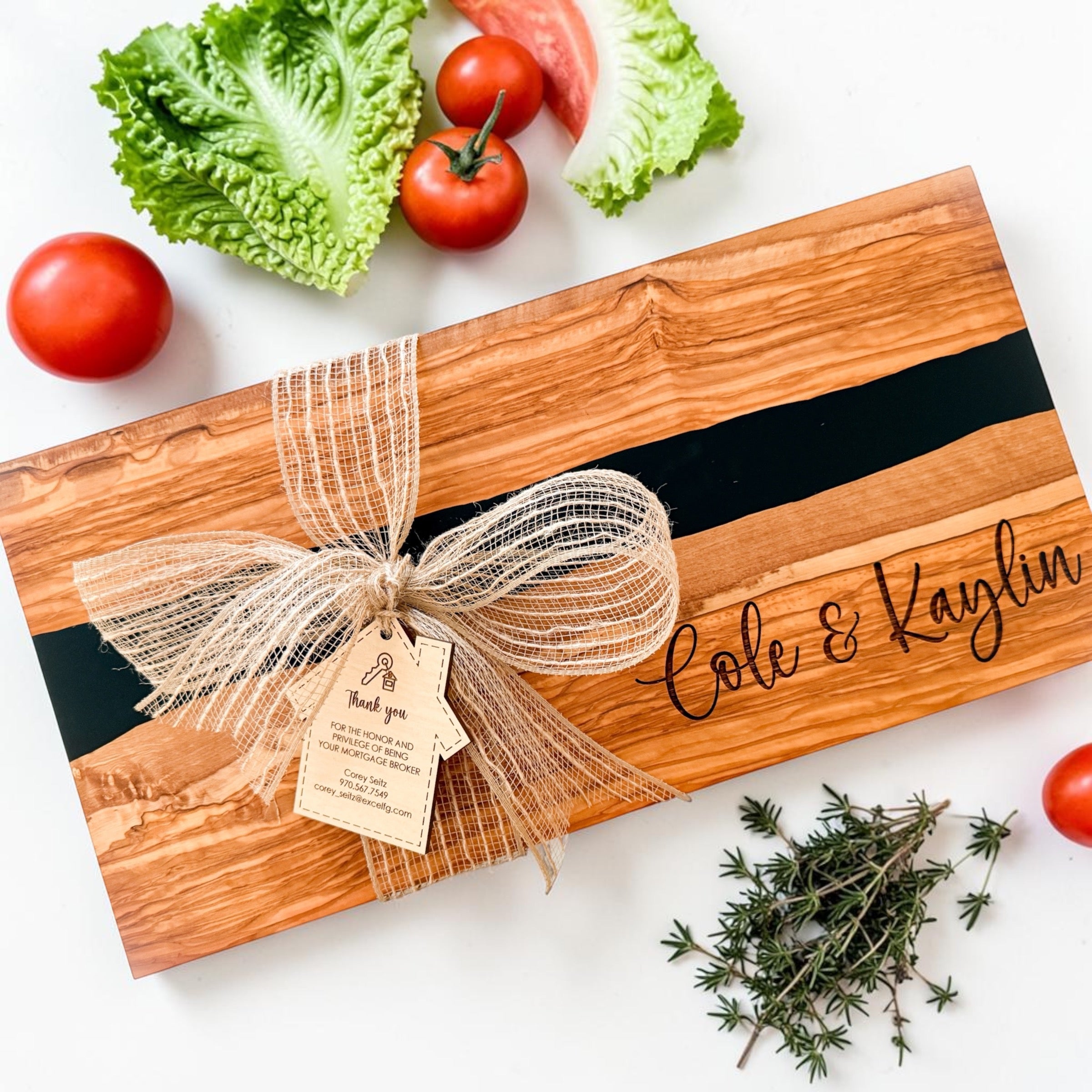 Personalized olive wood charcuterie board with black resin river detail, engraved “Cole & Kaylin” design, rustic burlap bow, and custom realtor thank-you tag styled with fresh tomatoes and herbs on a light neutral background. Luxury real estate closing gift and personalized kitchen gift for new homeowners.