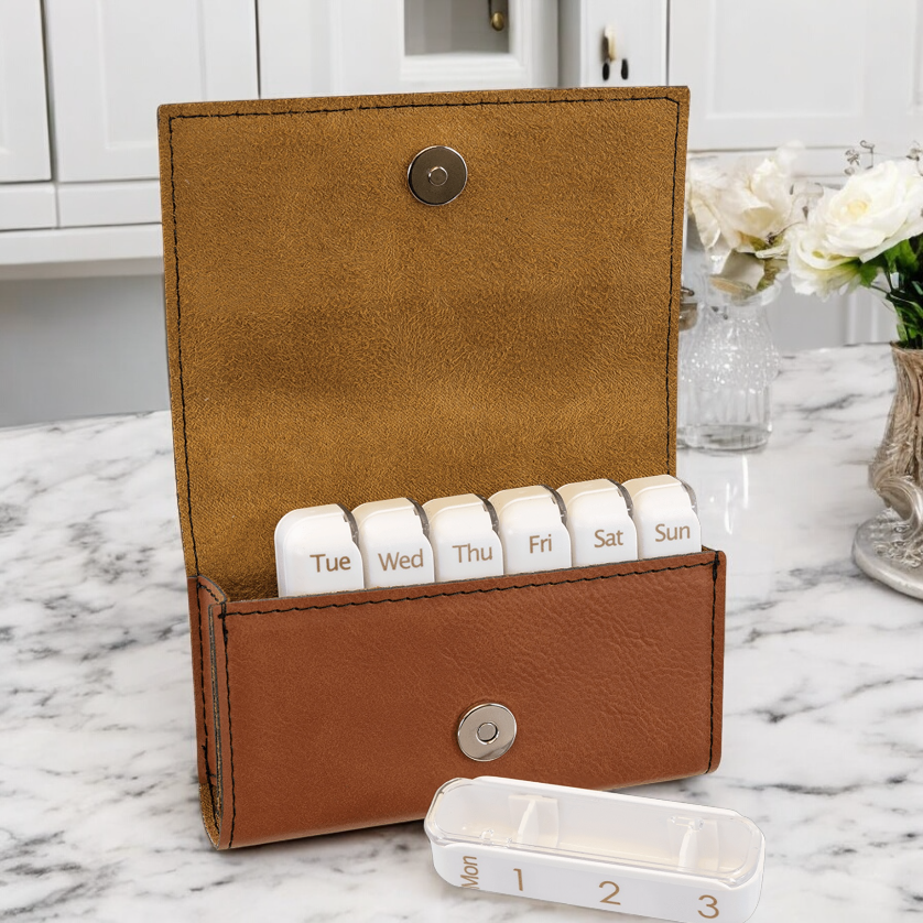 Custom laserable leatherette weekly pill organizer box featuring a compact design with a magnetic snap closure. Includes seven labeled plastic containers for daily pill organization, displayed in various colors such as black, blue, pink, and rawhide. Ideal for corporate branding, healthcare giveaways, or personal medication management.